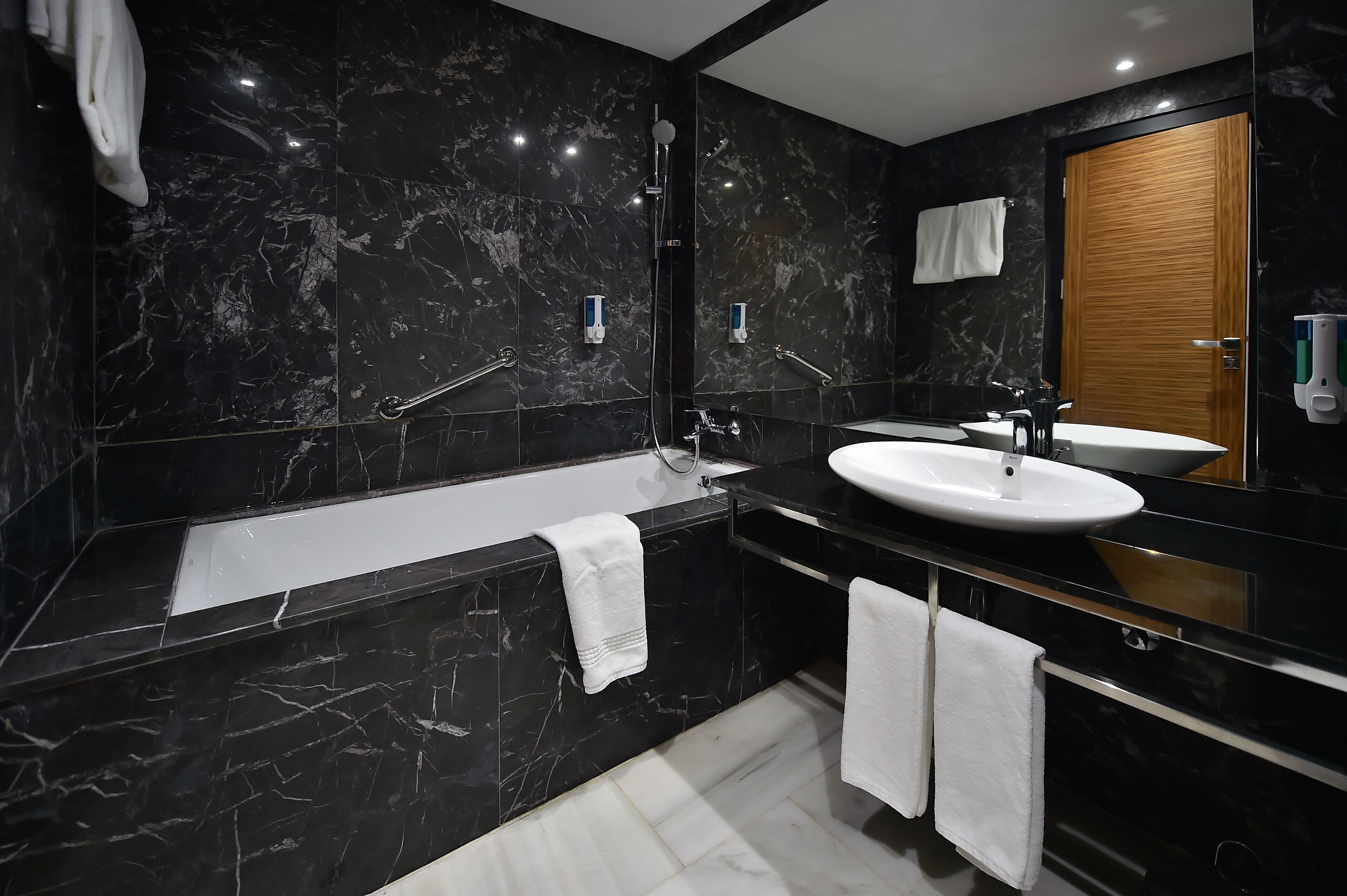 A modern bathroom with a black marble wall, a white bathtub, a sink, and a wooden door.
