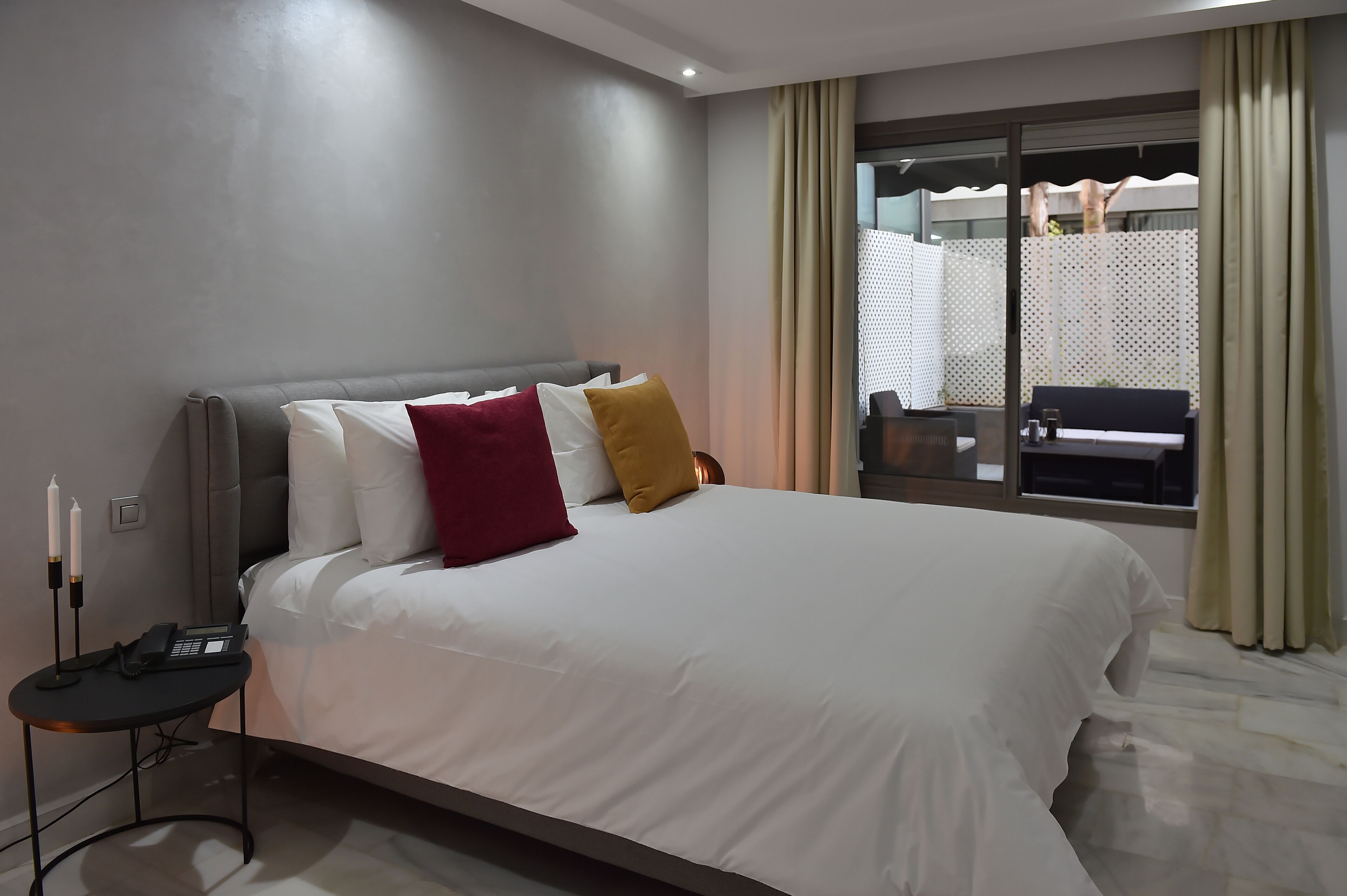 Quarto de hotel moderno com uma cama grande, uma mesinha de cabeceira com telefone e vista para a área externa através de uma janela.