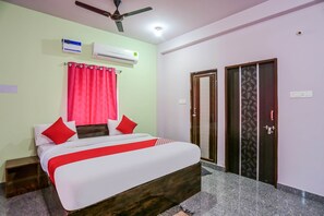 A hotel room with a bed, red curtains, an air conditioning unit, a ceiling fan, and a door with a decorative pattern.