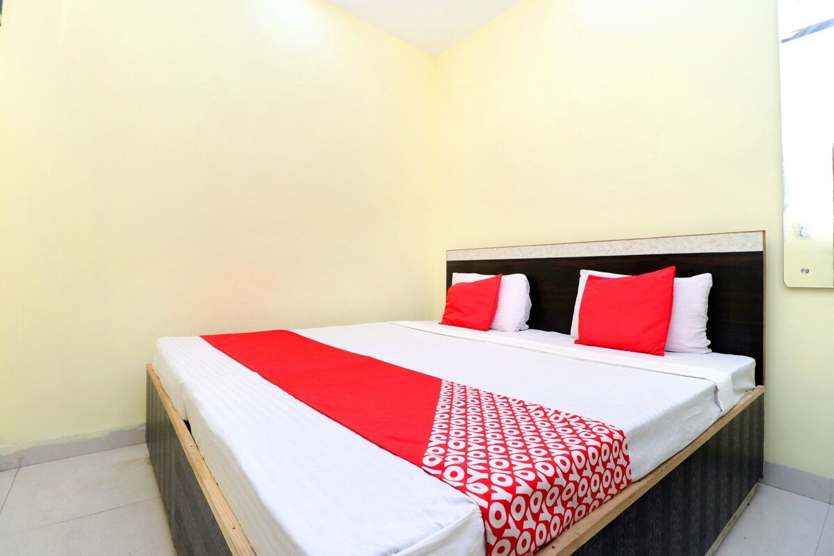 A single bed with a red and white patterned bedspread, two red pillows, and a wooden headboard in a room with light-colored walls and a tiled floor.