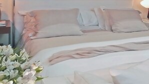 En pænt redt seng med hvide sengetæpper og et beige tæppe, et sengebord med en lampe, og en buket hvide blomster.