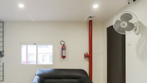 A room with a black leather sofa, a window, a fire extinguisher, a red pipe, and a white fan. - OYO Flagship 16390 Chukkapalli Chambers (Vijayawada)