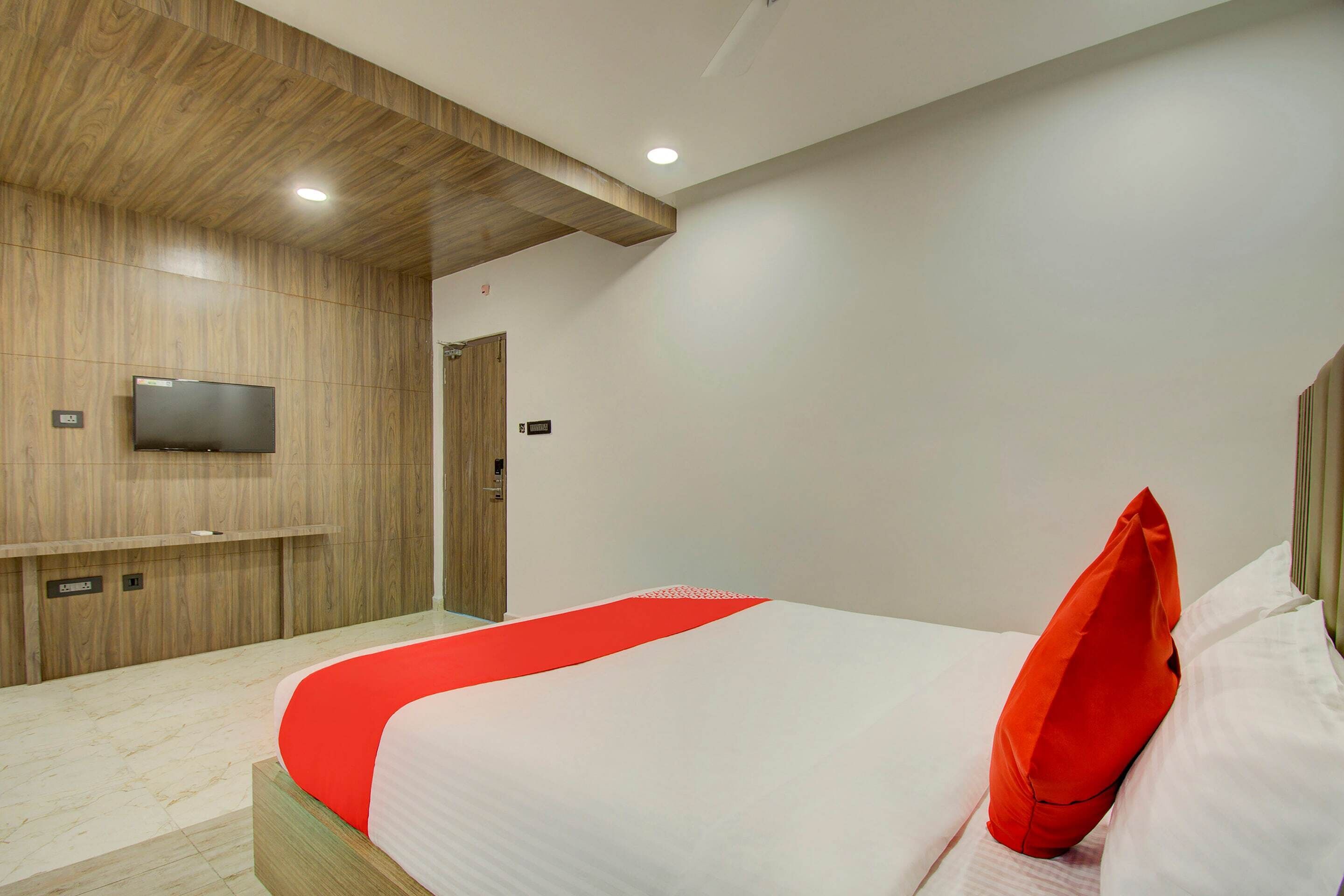A modern hotel room with a wooden accent wall, a flat-screen TV mounted above the bed, and a red accent pillow.