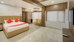 A modern hotel room with a wooden bed, two chairs, a TV, and a large window with blinds. - Capital O 23031 V Hotel (Penumuluru)