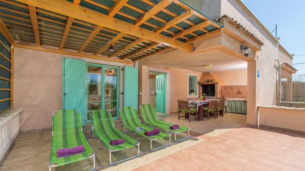 A covered outdoor area with four green lounge chairs, a dining table with chairs, and a kitchen area visible through an open door.