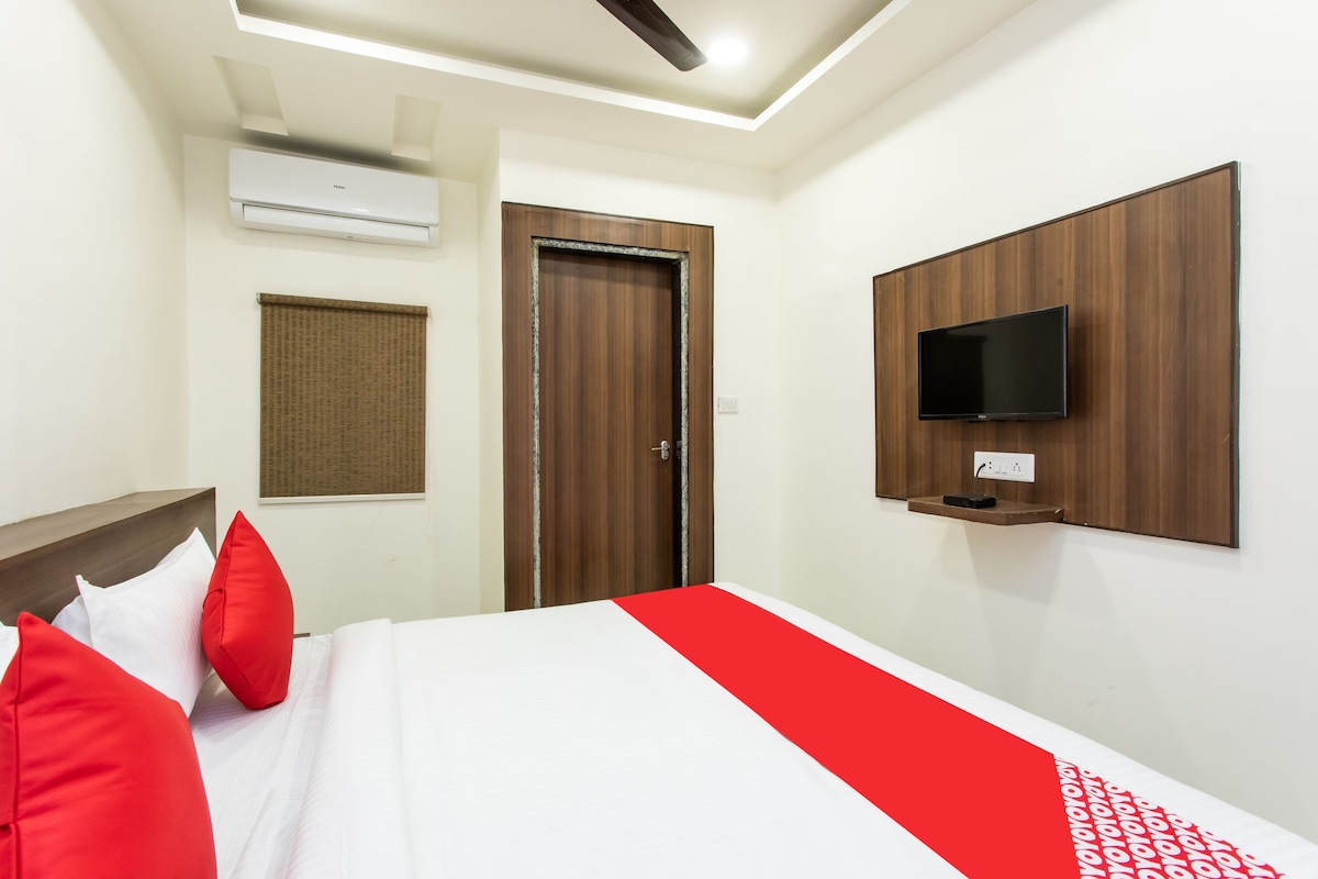 A hotel room with a bed, red pillows, a TV mounted on the wall, and an air conditioning unit.