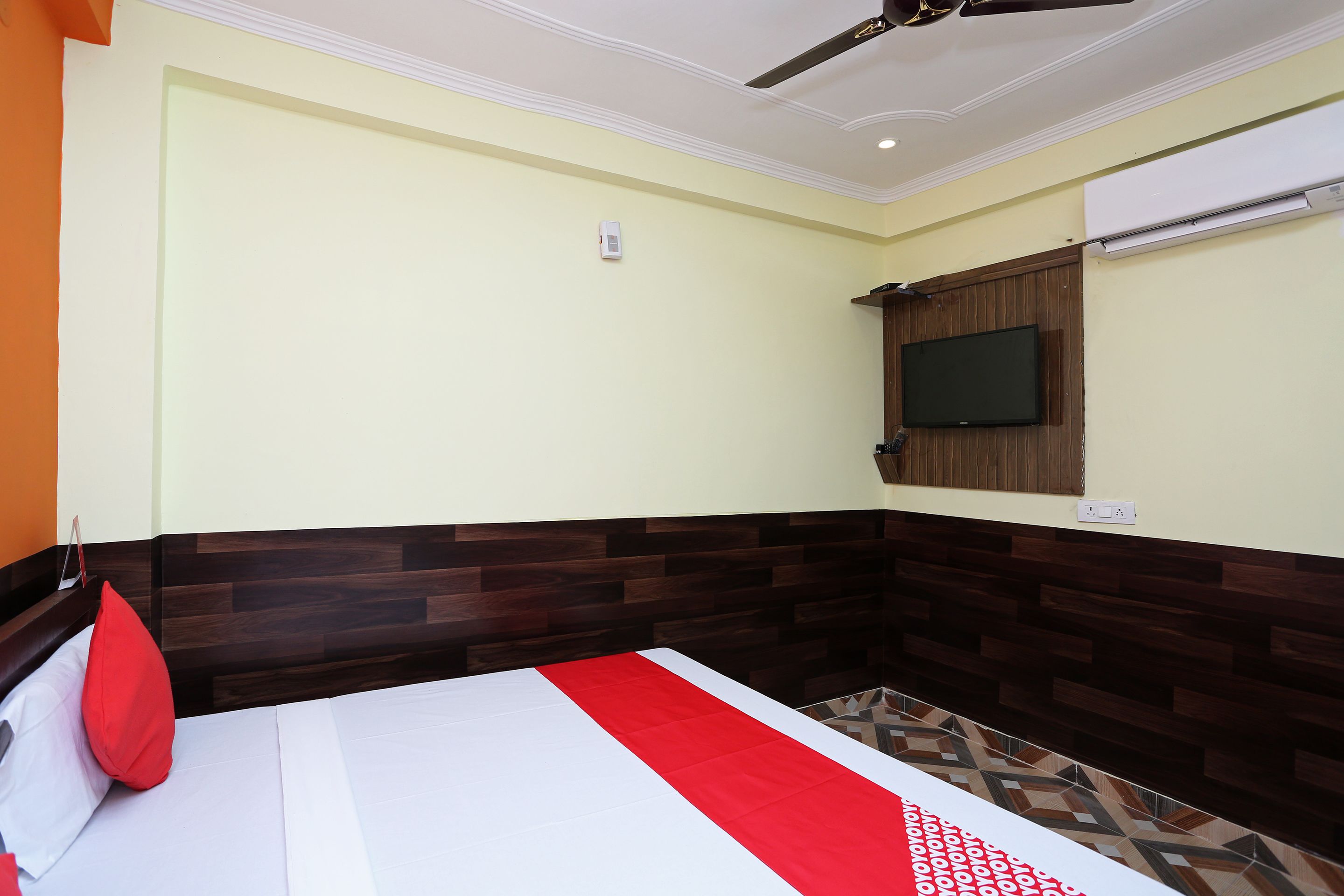 A hotel room with a bed, a red pillow, a ceiling fan, a mounted TV, and a patterned floor.