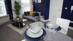 A modern living room with a glass coffee table, a sofa with colorful cushions, a dining table set with plates, glasses, and cutlery. - Cross St Boutique Apartment (Altrincham)