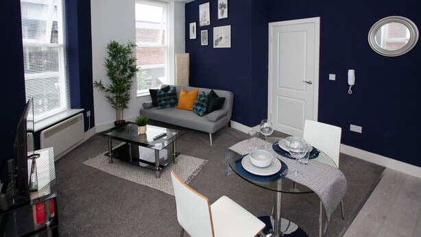 A modern living room with a grey sofa, a dining table with chairs, a large window with blinds, and a potted plant. - Cross St Boutique Apartment (Altrincham)