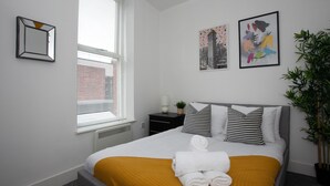 A neatly made bed with a yellow throw, a bedside table with a lamp, and a potted plant. - Cross St Boutique Apartment (Altrincham)