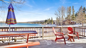 Una terraza de madera con una mesa de picnic y dos sillas Adirondack rojas, con vista a un lago y, al fondo, árboles y edificios.