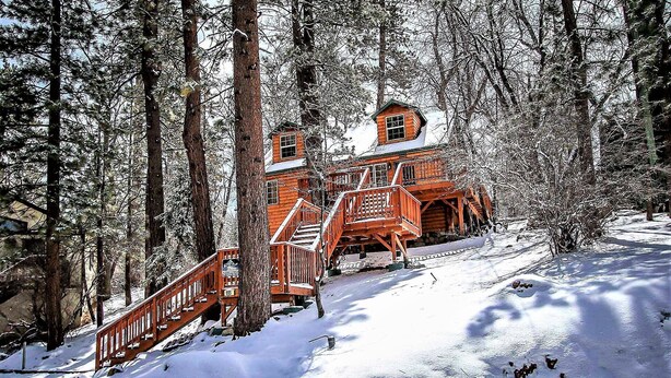 A wooden cabin with a staircase leading up to a deck, surrounded by snow-covered trees. - Nine Pines Lodge#537 (Big Bear City)