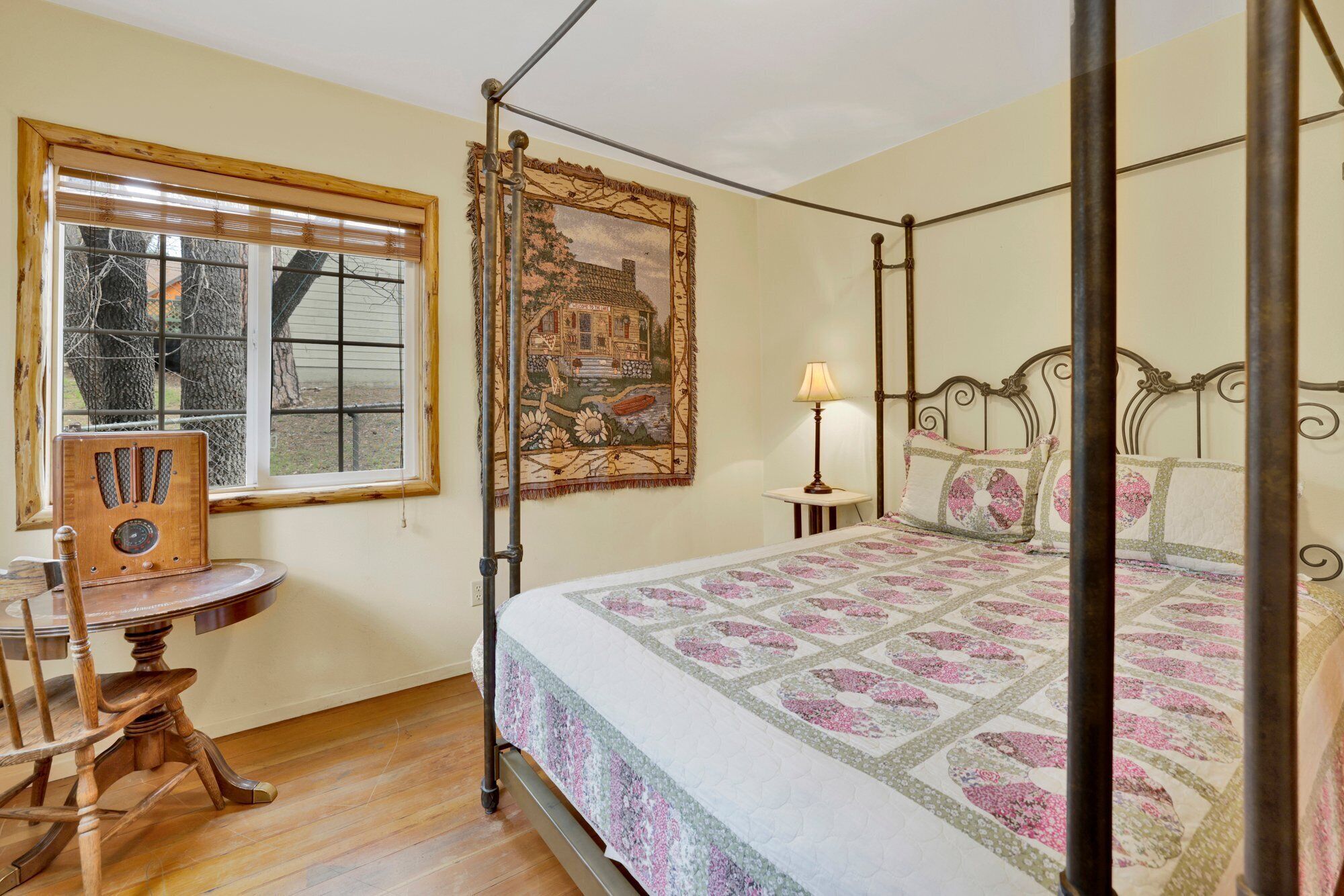 A four-poster bed with a floral quilt, a wooden rocking chair, a vintage radio, and a large landscape painting on the wall.