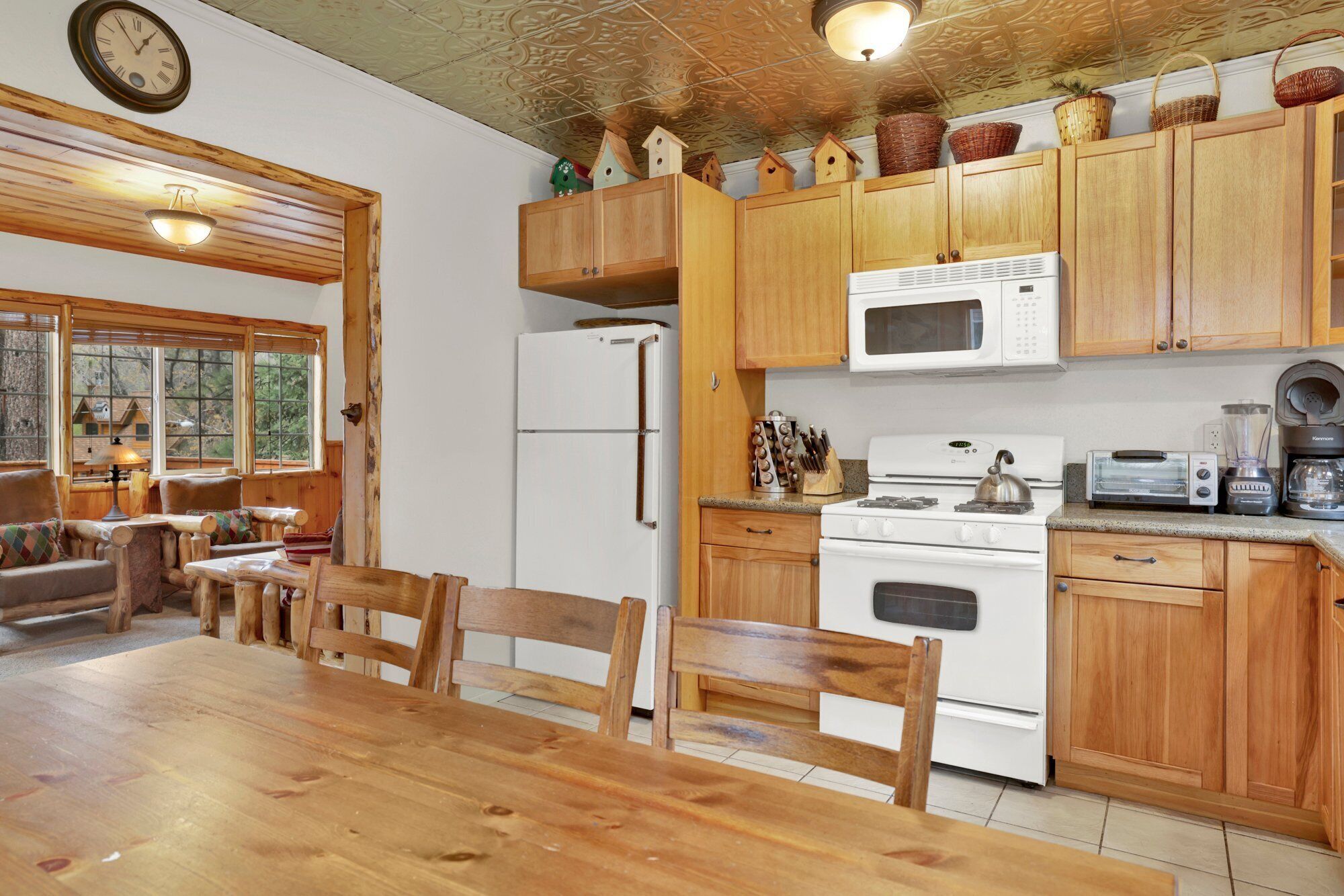 A rustic kitchen with wooden cabinets, a white refrigerator, a stove, and a microwave. There is a dining table with chairs and a window with a view of trees.