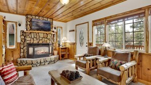 A cozy cabin interior with a stone fireplace, wooden furniture, and large windows offering a view of the outdoors. - Nine Pines Lodge#537 (Big Bear City)