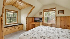 A bedroom with a sloped ceiling, wooden walls, a bed with a patterned bedspread, a desk with a television, and two windows with views of trees. - Nine Pines Lodge#537 (Big Bear City)