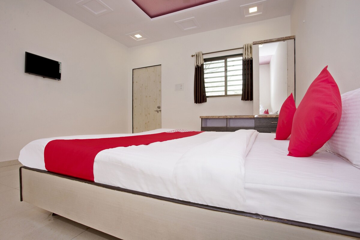 A bedroom with a bed, a mirror, a window with curtains, and a flat-screen TV mounted on the wall.