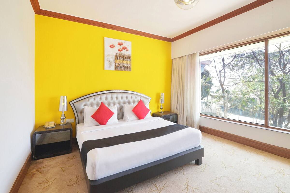 A hotel room with a bed, two bedside tables, a telephone, a painting, and a large window with a view of trees.