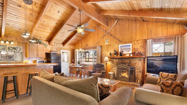 A cozy log cabin interior with a dining area, a fireplace, and a large window overlooking a scenic view. - Oriole Cottage #621 (Big Bear Lake)