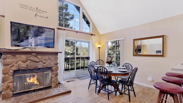 A cozy living room with a stone fireplace, a dining table with chairs, and a large window overlooking a deck. - Bear Mountain Chalet #639 (Big Bear Lake)