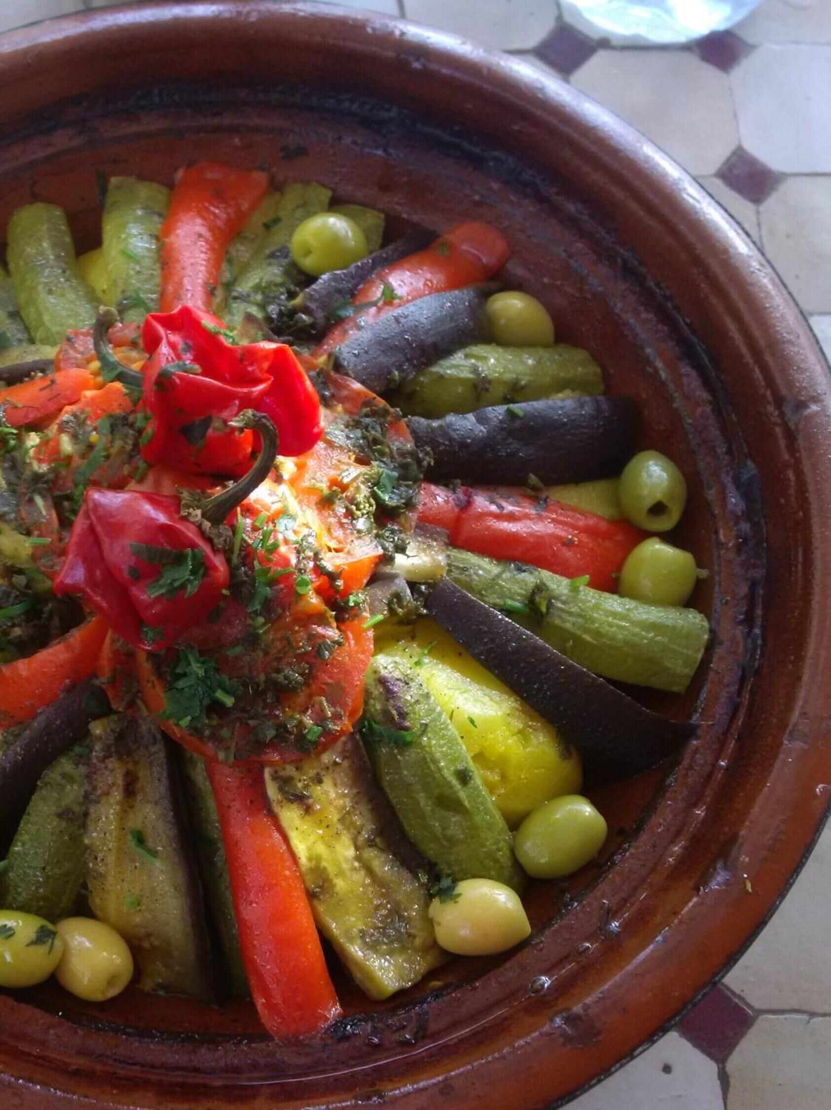 A terracotta dish filled with roasted vegetables including carrots, eggplants, and olives, garnished with herbs.