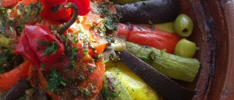 A terracotta dish filled with roasted vegetables including carrots, eggplants, and olives, garnished with herbs.