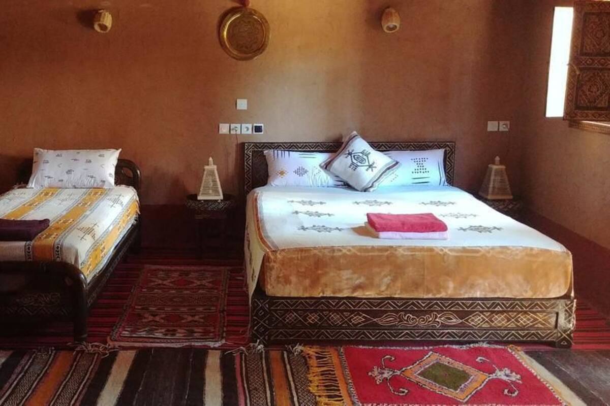 Een kamer met twee bedden, elk voorzien van beddengoed en kussens met een patroon, een zwart-rood gestreept tapijt en een klein tafeltje met een lamp.