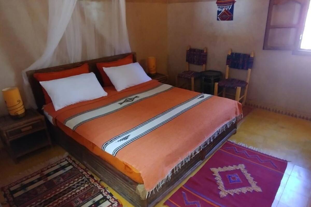 Een bed met hemel, twee kussens, een geborduurde deken en een nachtkastje met een lamp.