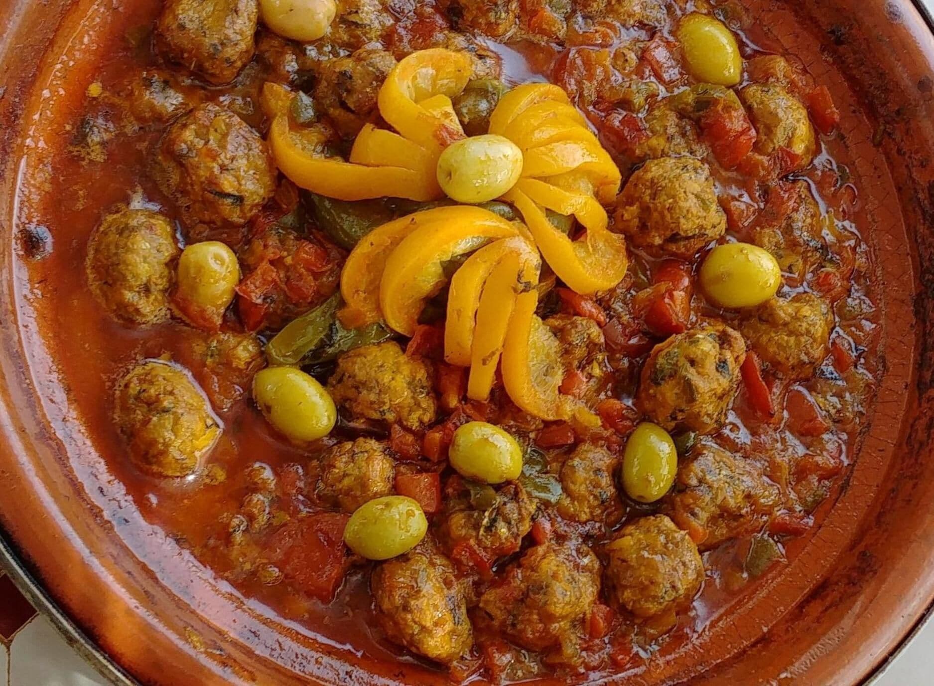 A large ceramic dish filled with meatballs, yellow bell peppers, and olives in a tomato-based sauce.