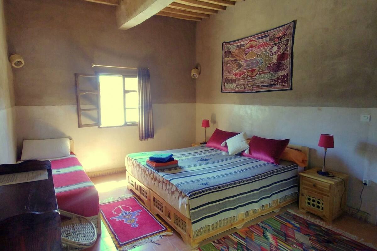 Een slaapkamer met een bed, kussens, een nachtkastje, een lamp, een raam met gordijnen, een wanddecoratie en een kamerbreed tapijt.