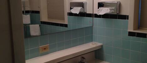 Une salle de bain avec un lavabo, un miroir et des toilettes. Il y a des lampes au-dessus du miroir et un distributeur de serviettes.