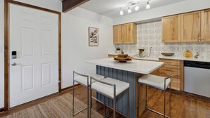 Una cocina moderna con armarios de madera, encimera blanca y lavavajillas integrado. Hay dos taburetes de bar y una pequeña isla con un bol encima.