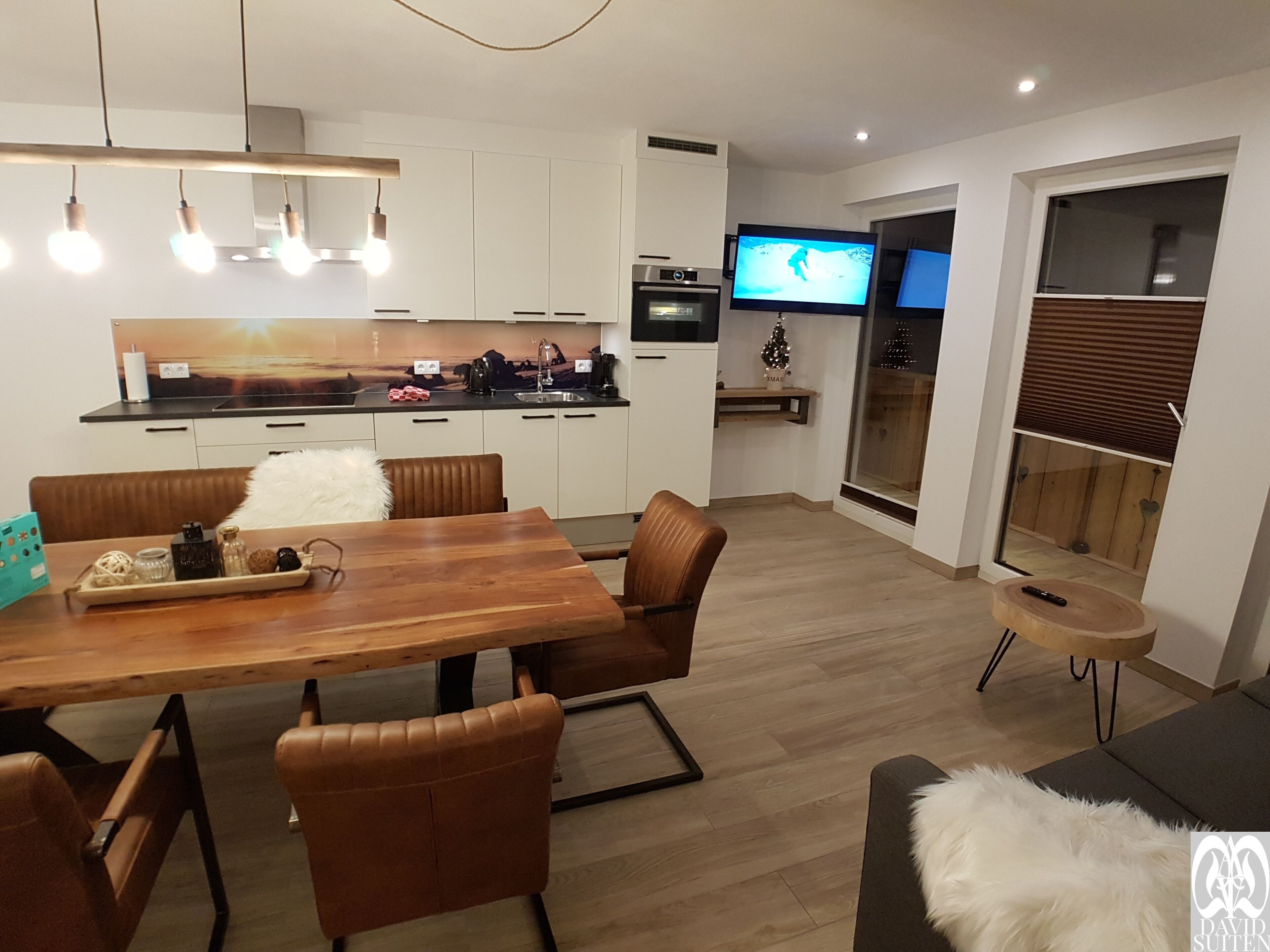 A modern kitchen with white cabinets, a wooden dining table, and a TV mounted on the wall.