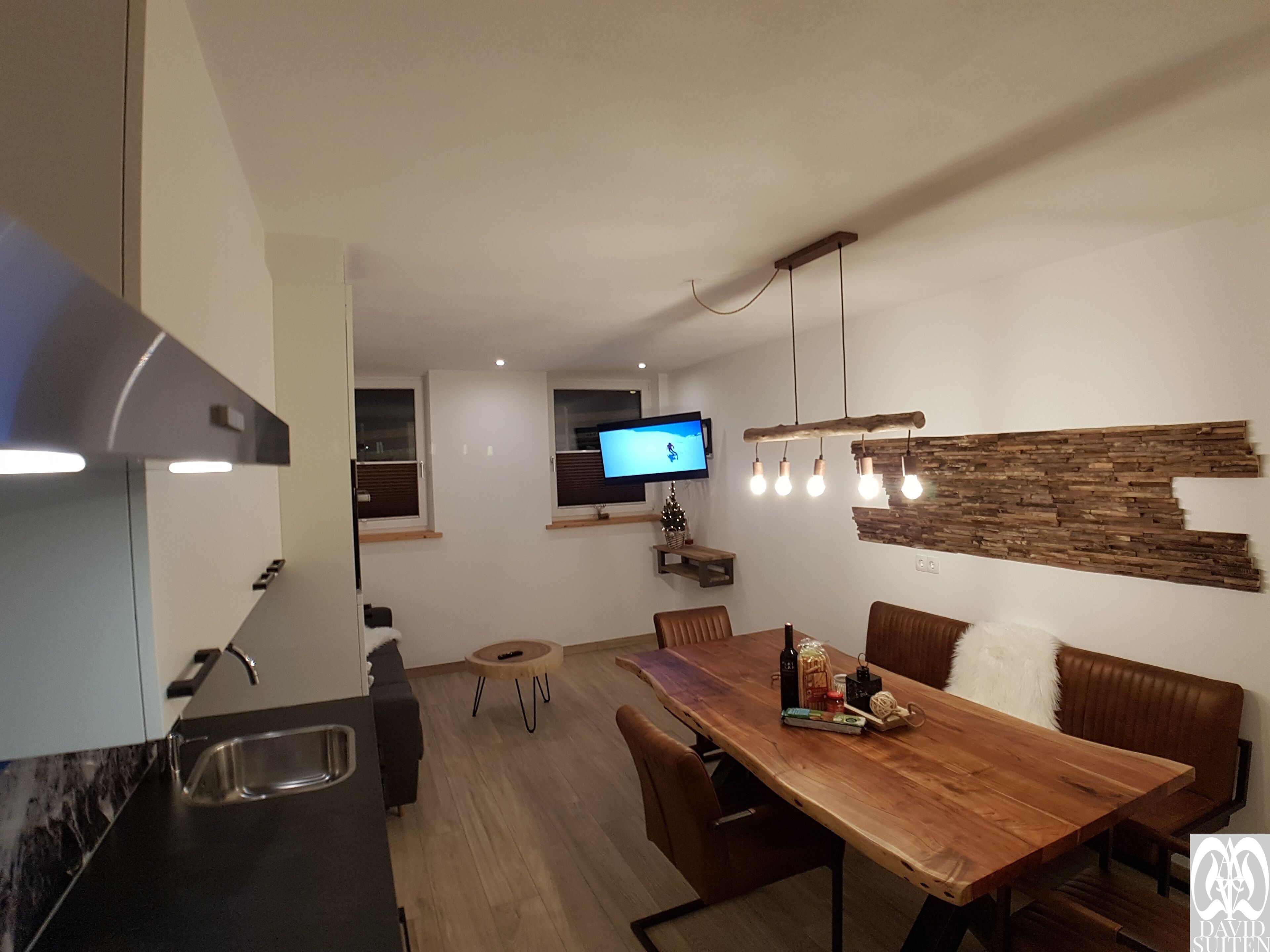 A modern kitchen with a dining area, a TV mounted on the wall, and a wooden table with chairs.