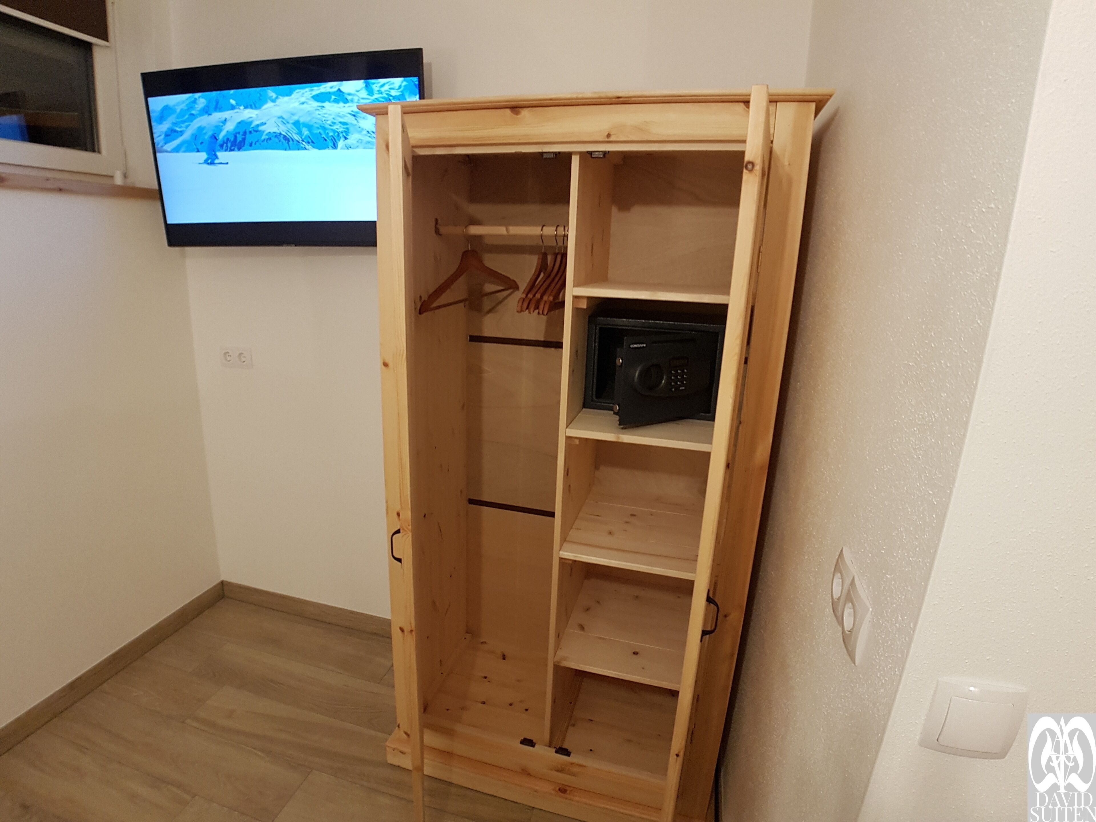 Um guarda-roupa de madeira com várias prateleiras e uma barra para pendurar roupas, uma televisão fixada na parede e uma tomada elétrica na parede.