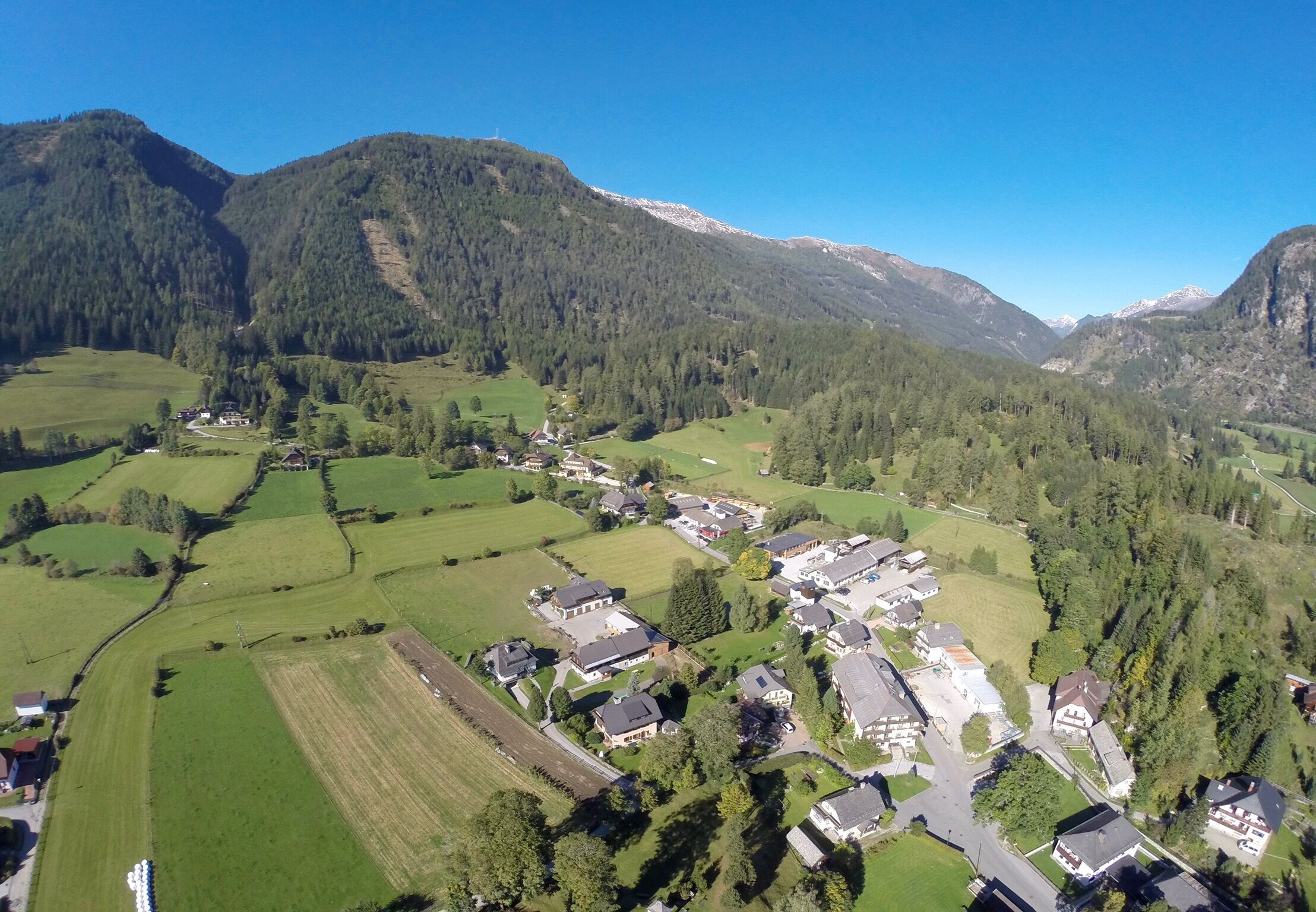 Eine Vogelperspektive auf ein Dorf mit Häusern, grünen Feldern und Bergen im Hintergrund.
