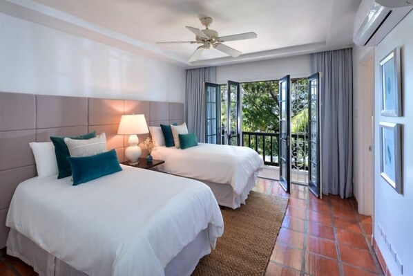 A hotel room with two beds, a ceiling fan, a nightstand with a lamp, and a view of the outdoors through a glass door.
