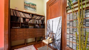 A room with a wooden bookshelf filled with records, a chair, a large painting of a cityscape, and a potted plant.