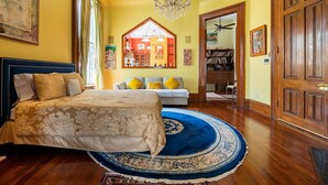 A bedroom with a large bed, a blue and white patterned rug, wooden flooring, a chandelier, and a view of a room with a ceiling fan.