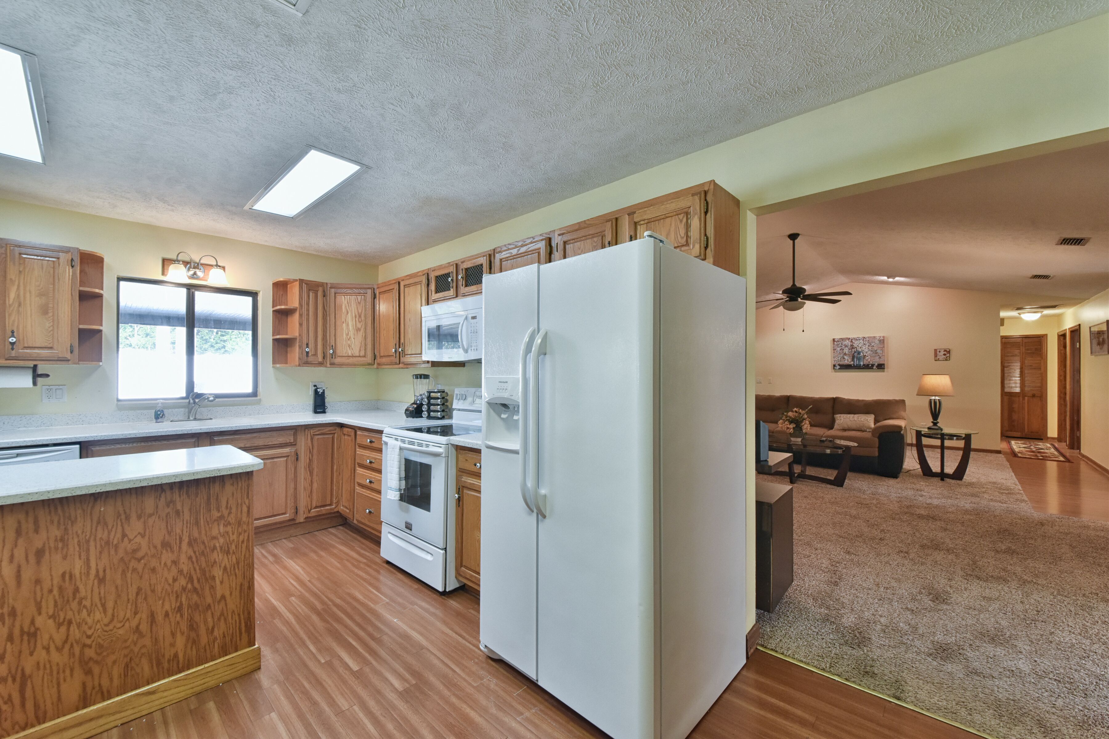 A kitchen with wooden cabinets, a white refrigerator, a microwave, and an oven. There is a window with a view of trees, and a ceiling fan.