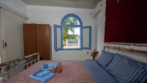 Un dormitorio con cama, armario de madera, ventana con contraventanas azules, cortina roja y una pequeña mesa con un jarrón de flores.