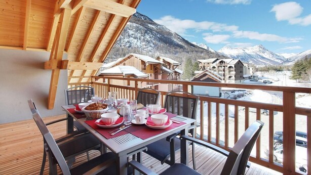 Eine Holzterrasse mit einem für das Frühstück gedeckten Tisch, mit Blick auf eine verschneite Berglandschaft.