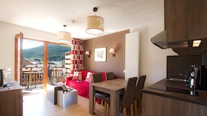 Ein modernes Wohnzimmer mit einem roten Sofa, einem Holzespeisetisch und Stühlen, einer Küchenzeile mit einer Kaffeemaschine und einem Balkon mit Bergblick.