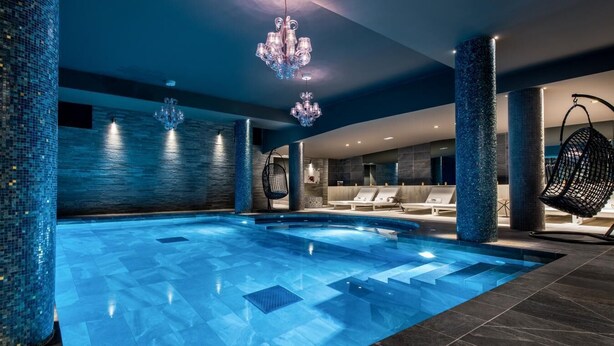 A modern indoor swimming pool with a blue mosaic tiled floor, a chandelier, and lounge chairs.