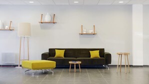 Un salon moderne avec un canapé vert foncé, deux coussins jaunes, un pouf jaune, une lampe sur pied et deux petites tables.