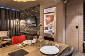 Quarto de hotel moderno com uma mesa de jantar de madeira posta para dois, um espaço de estar aconchegante com uma poltrona vermelha, uma grande TV de tela plana fixada em uma parede de pedra, e um quarto visível com uma cama e uma mesinha de cabeceira.