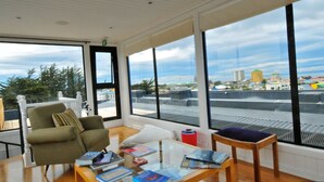 A room with a large window, a green armchair, a small table with books, and a view of buildings and trees. - Hotel Ilaia (Punta Arenas)