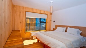 A bedroom with a wooden floor, a large window with a view, a bed with white bedding, and a wooden headboard. - Hotel Ilaia (Punta Arenas)