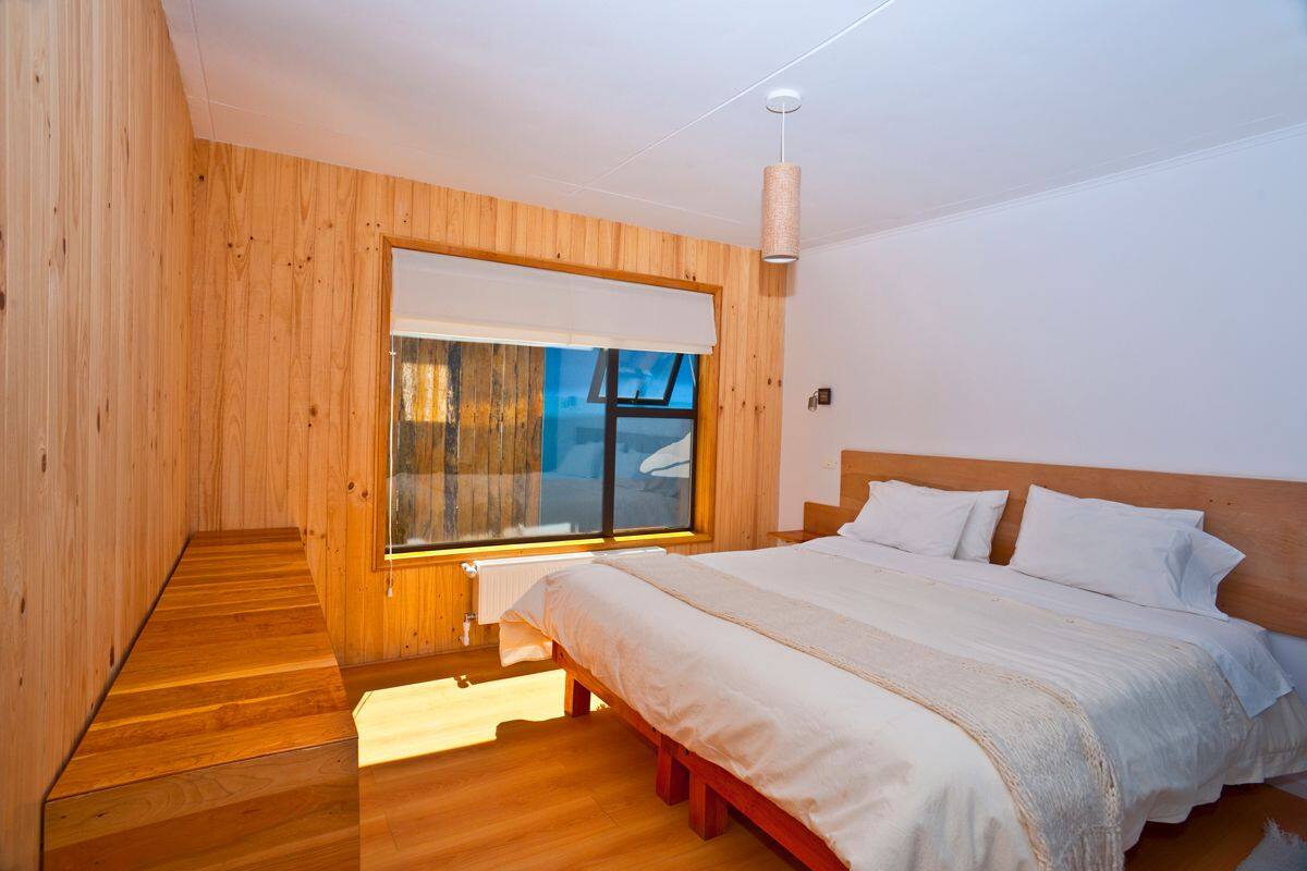 A bedroom with a wooden floor, a large window with a view, a bed with white bedding, and a wooden headboard.