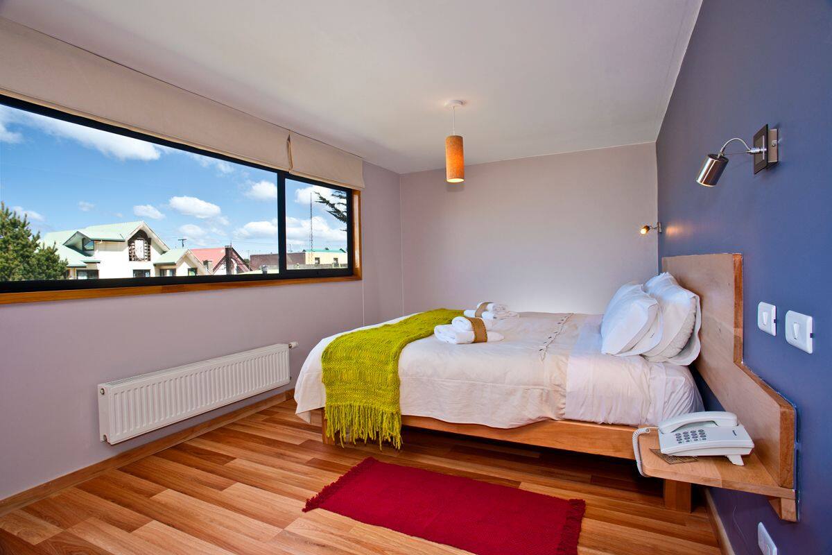 A modern bedroom with a large window, a bed with pillows and a blanket, a bedside table with a phone, and a red rug on the floor.